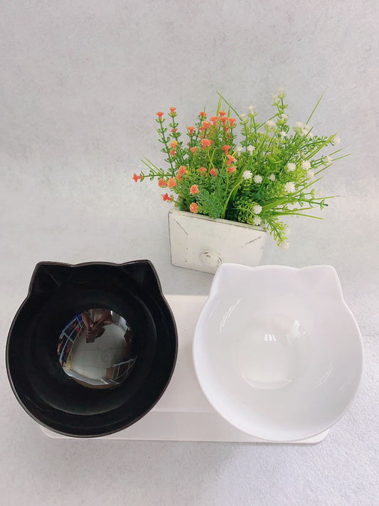 Two cat-shaped bowls, one black and one white, with a small potted plant on a light gray background.